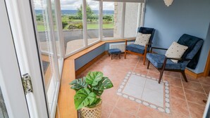 A sunroom with a view of the countryside, a seating area with two chairs and a bench, and a potted plant. - Braeside Farm House (Ballymena)