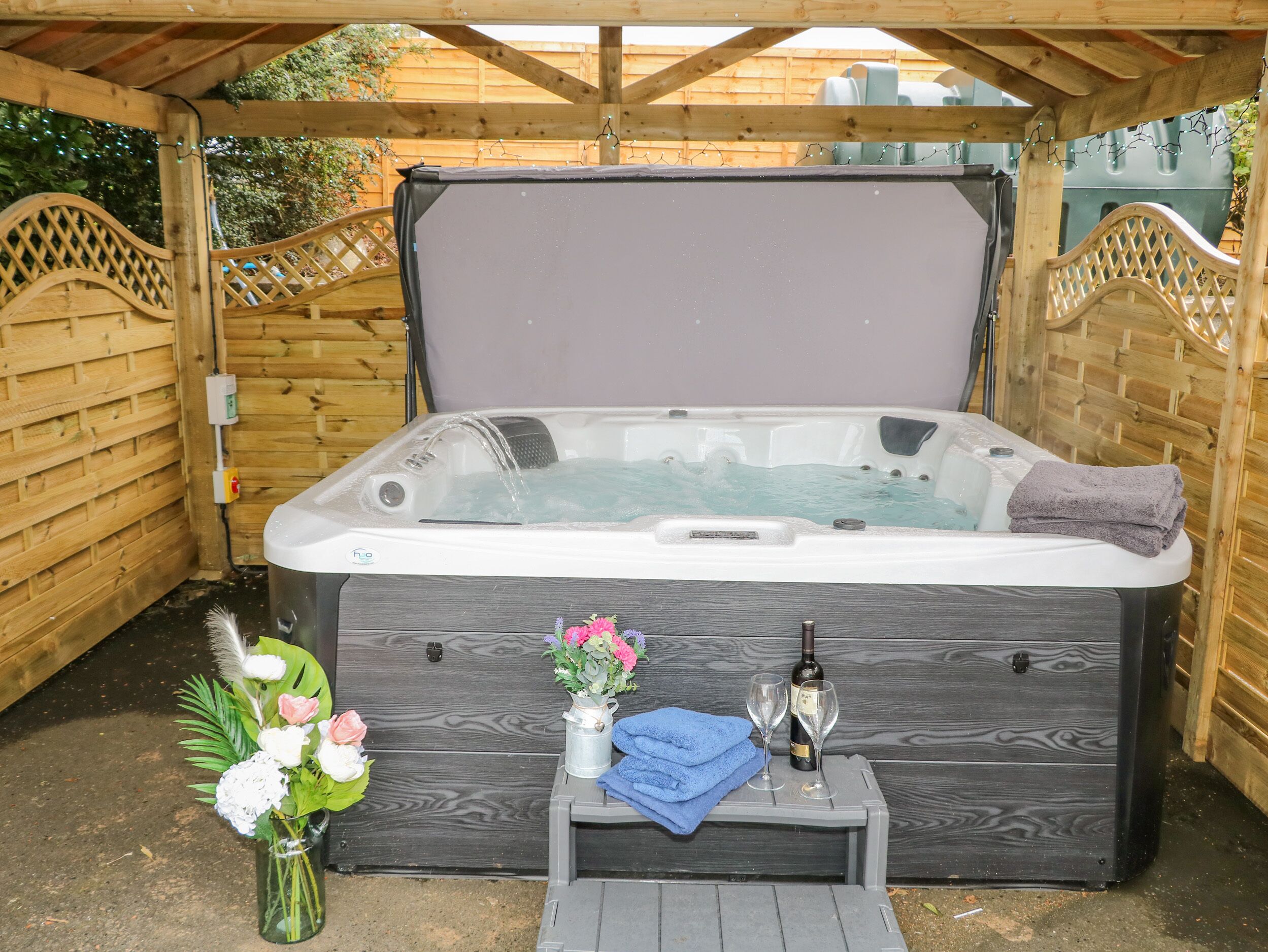 A hot tub with a wooden deck, a small table with a bottle and glasses, and a vase of flowers.