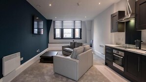 Een moderne woonkamer met een grijze bank en een salontafel, een keuken met donkere kasten en een roestvrijstalen oven, en grote ramen met zonwering.