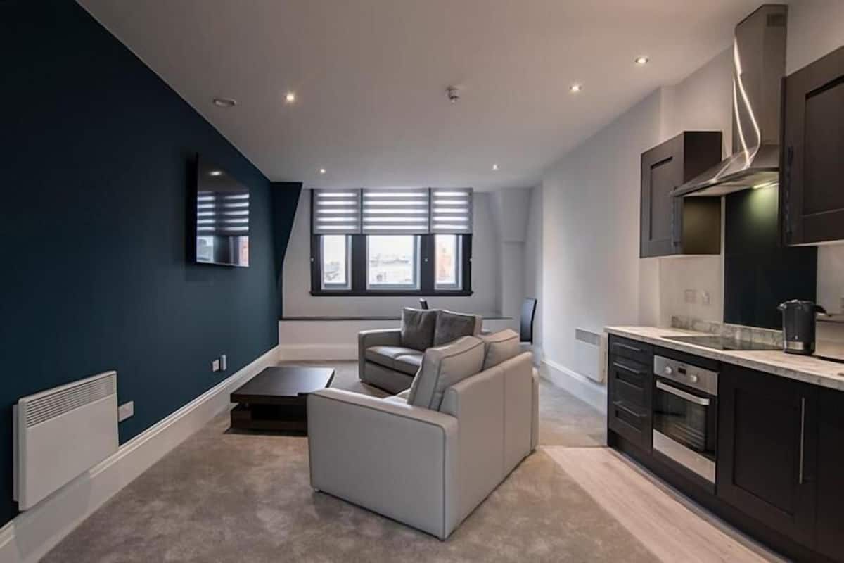 Ein modernes Wohnzimmer mit grauem Sofa und Couchtisch, eine Küche mit dunklen Schränken und einem Edelstahlbackofen sowie große Fenster mit Jalousien.