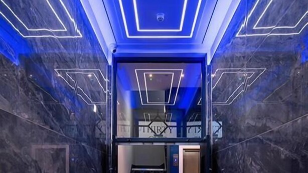 Een moderne liftopgang met een geometrisch lichtontwerp in het plafond en marmeren wanden.