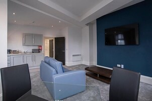 Una sala de estar moderna con un sofá azul, sillas negras, un televisor de pantalla plana sobre una pared azul y una cocina al fondo.