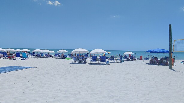 A beach with rows of blue chairs and white umbrellas, a volleyball net, and a sandy shore.