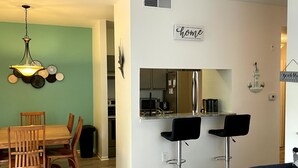 A modern kitchen with a stainless steel refrigerator, a microwave, and a coffee maker. There is a dining area with a wooden table and chairs, and a bar area with two black bar stools.