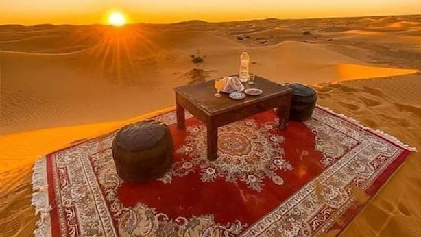 A desert scene with a table set for two, a rug, and a tent in the background. - Camel Trek Bivouac in Merzouga (Taouz)