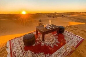 A desert scene with a table set for two, a rug, and a tent in the background. - Camel Trek Bivouac in Merzouga (Taouz)