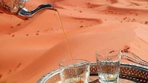 A tray with two glasses, one with a dark liquid and the other with a clear liquid, and a metal pitcher pouring a golden liquid into the clear glass. - Camel Trek Bivouac in Merzouga (Taouz)