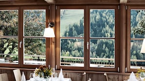 A dining area with wooden paneled walls and large windows offering a view of a forested landscape. Tables are set with white tablecloths, napkins, and glassware. - Naturhotel Saalerwirt (San Lorenzo di Sebato)
