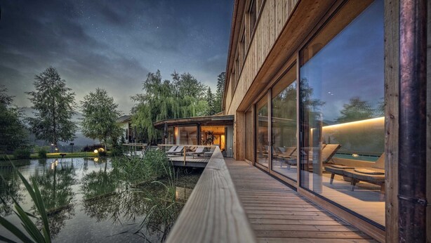 A modern building with large glass windows, a wooden deck, and a view of a lake at night. - Naturhotel Saalerwirt (San Lorenzo di Sebato)