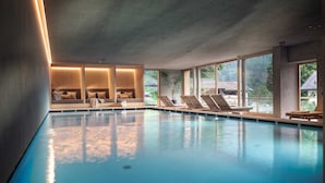 A modern indoor swimming pool with clear glass walls, lounge chairs, and a seating area with cushions. - Naturhotel Saalerwirt (San Lorenzo di Sebato)