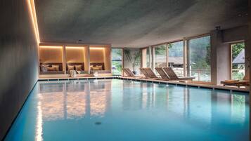 A modern indoor swimming pool with clear glass walls, lounge chairs, and a seating area with cushions.