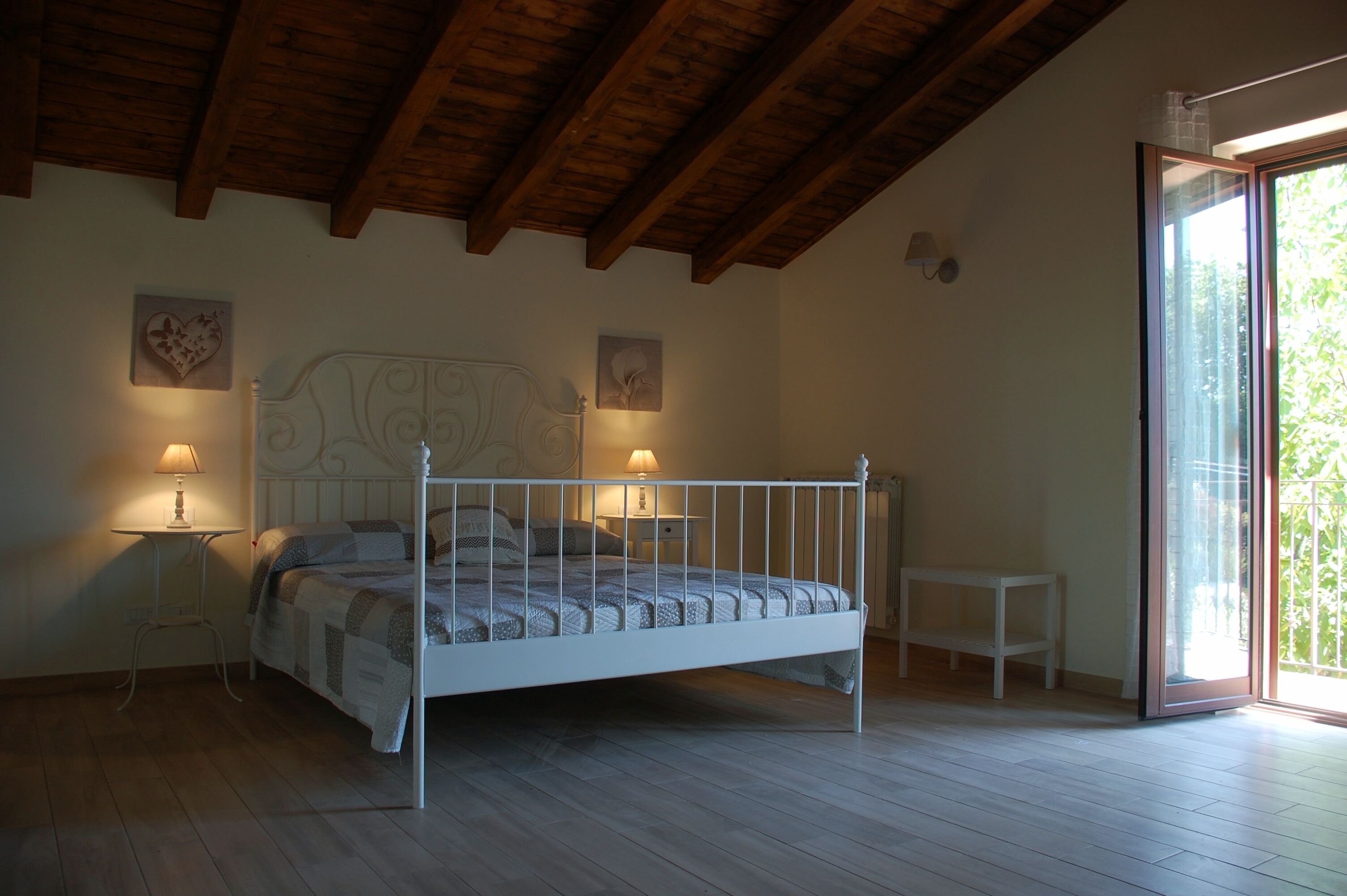 Ein Schlafzimmer mit einem weißen Metallbett, zwei Nachttischen mit Lampen, einem kleinen weißen Tisch und Blick auf Grün durch eine Schiebetür.
