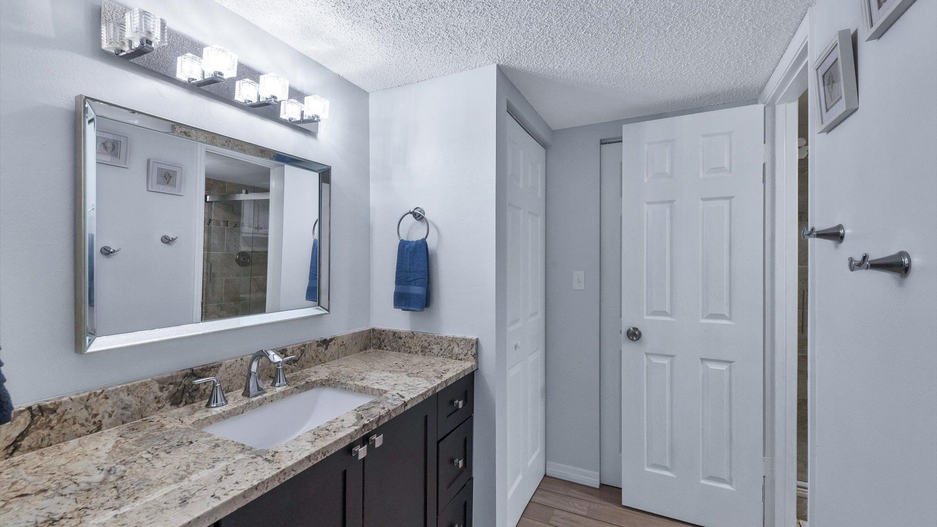 Un baño con un espejo grande, un lavamanos y una ducha. Hay un perchero con una toalla azul colgada. Las paredes están pintadas de blanco y hay una puerta con un tirador plateado.