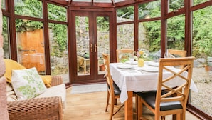 A conservatory with a wooden table set for dining, surrounded by wicker furniture and a stone wall. - The Lodge (Penrith)