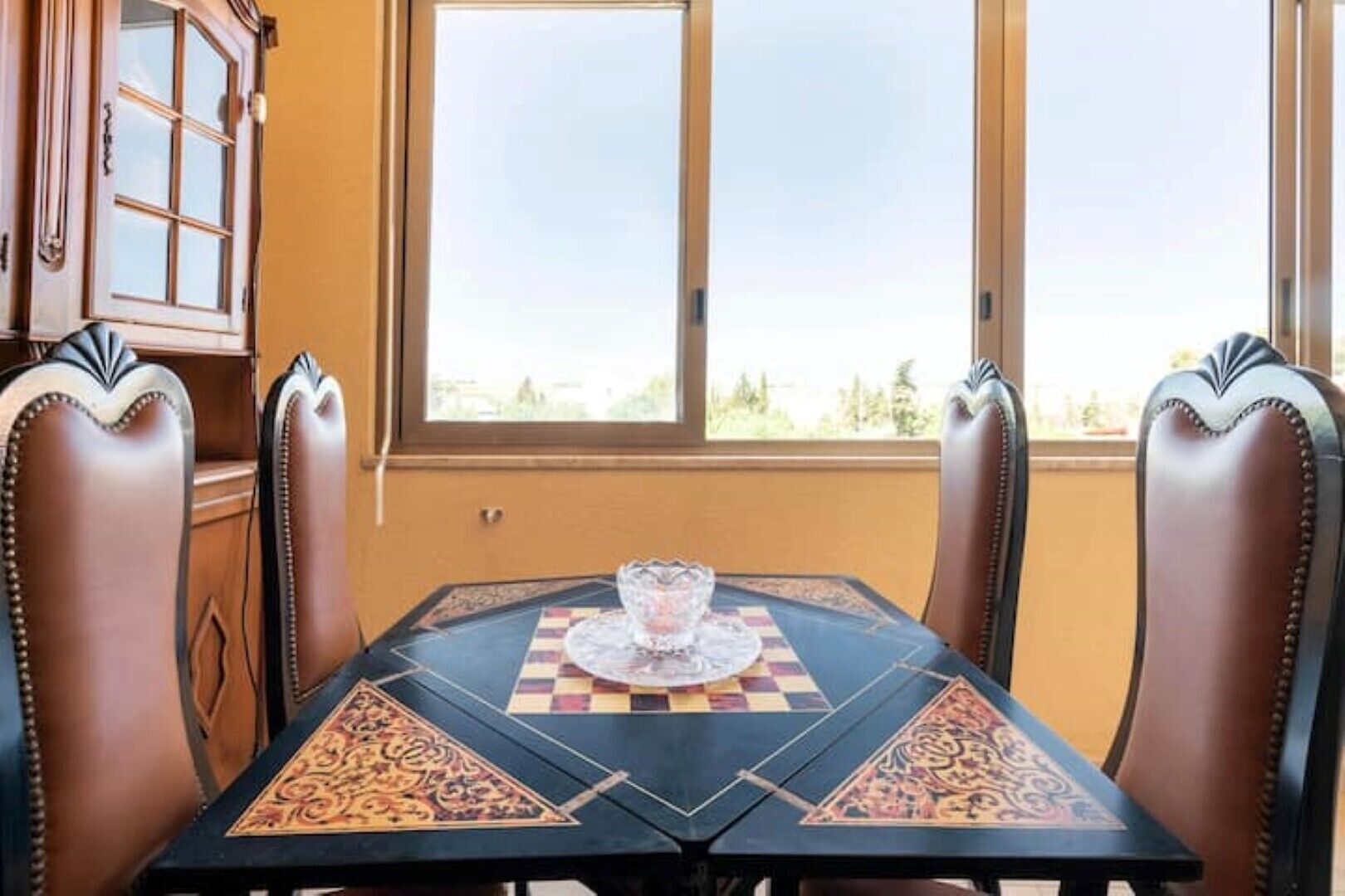 Un coin repas avec une table dressée pour deux personnes, agrémenté d’un jeu d’échecs en guise de décoration centrale, de fauteuils en cuir et offrant une vue à travers une grande fenêtre.