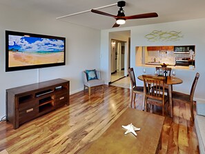 Un vaste espace de vie doté d’un plancher en bois, d’un téléviseur à écran plat fixé au mur, d’une table à manger avec des chaises, d’un ventilateur de plafond et d’un coin cuisine visible à travers une porte ouverte.