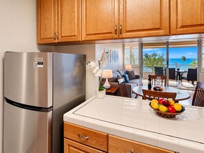 A kitchen with a stainless steel refrigerator, wooden cabinets, a dining area with a table and chairs, and a view of the ocean through large windows. - Prime Kihei 2BR Condo Breathtaking Panoramic Ocean Views Kamaole I Beach (Kihei)