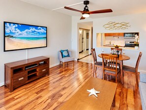 A modern living space with a flat-screen TV, wooden entertainment unit, dining table with chairs, and a kitchen area. - Prime Kihei 2BR Condo Breathtaking Panoramic Ocean Views Kamaole I Beach (Kihei)
