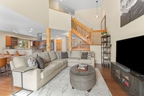 A spacious living area with a large sectional sofa, ottoman, and a flat-screen TV. A wooden staircase connects the living area to the kitchen.