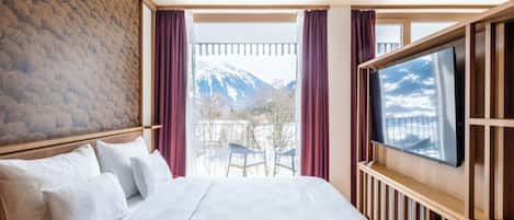 A hotel room with a large bed, a TV mounted on the wall, and a balcony with a mountain view.