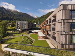 A modern building with multiple levels and balconies, surrounded by a well-maintained garden and a mountainous backdrop. - Falkensteiner Family Hotel Montafon - The Leading Hotels of the World (Tschagguns)