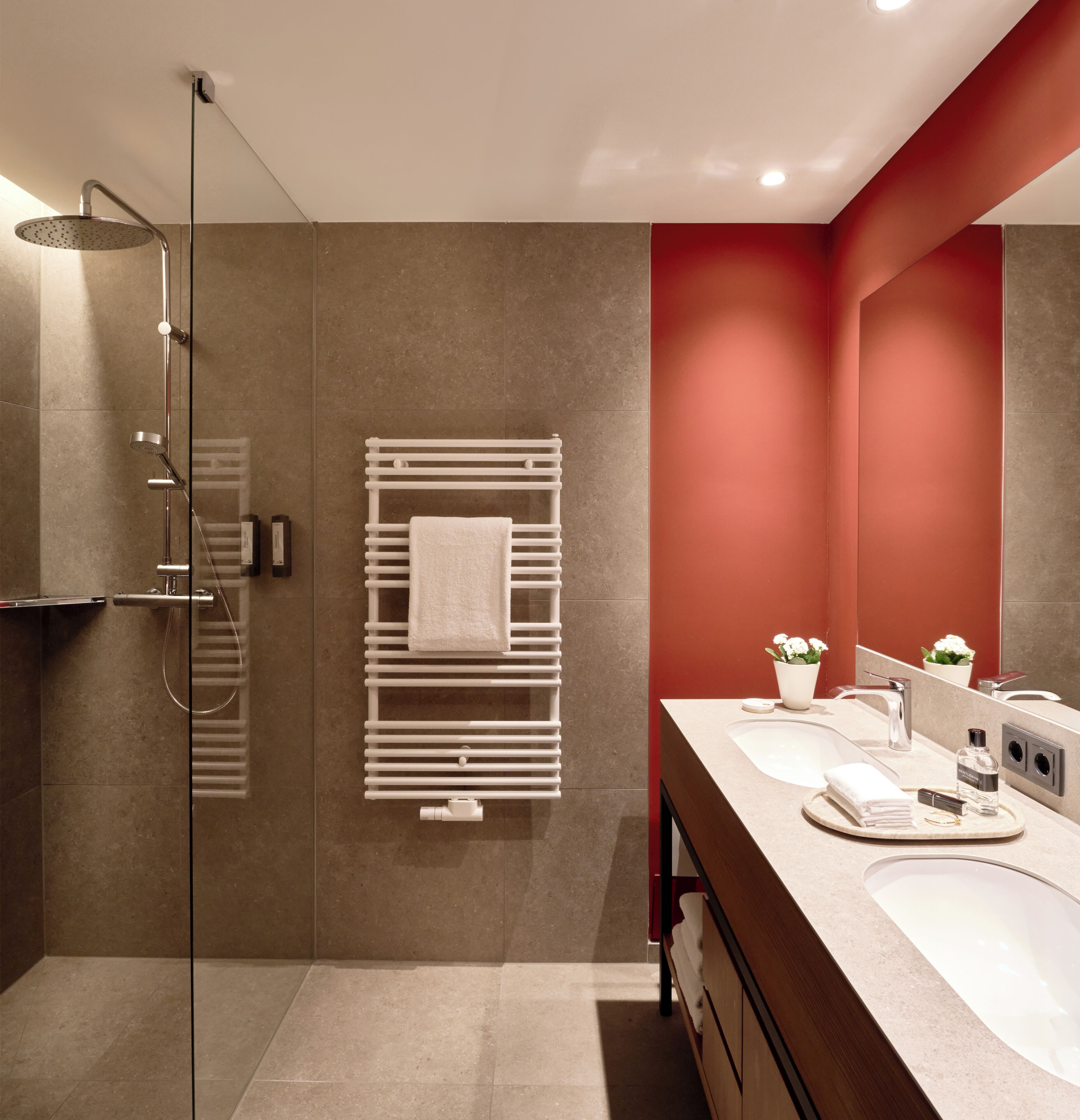 A modern bathroom with a glass shower enclosure, a white towel rack, a red accent wall, and a vanity with a mirror.