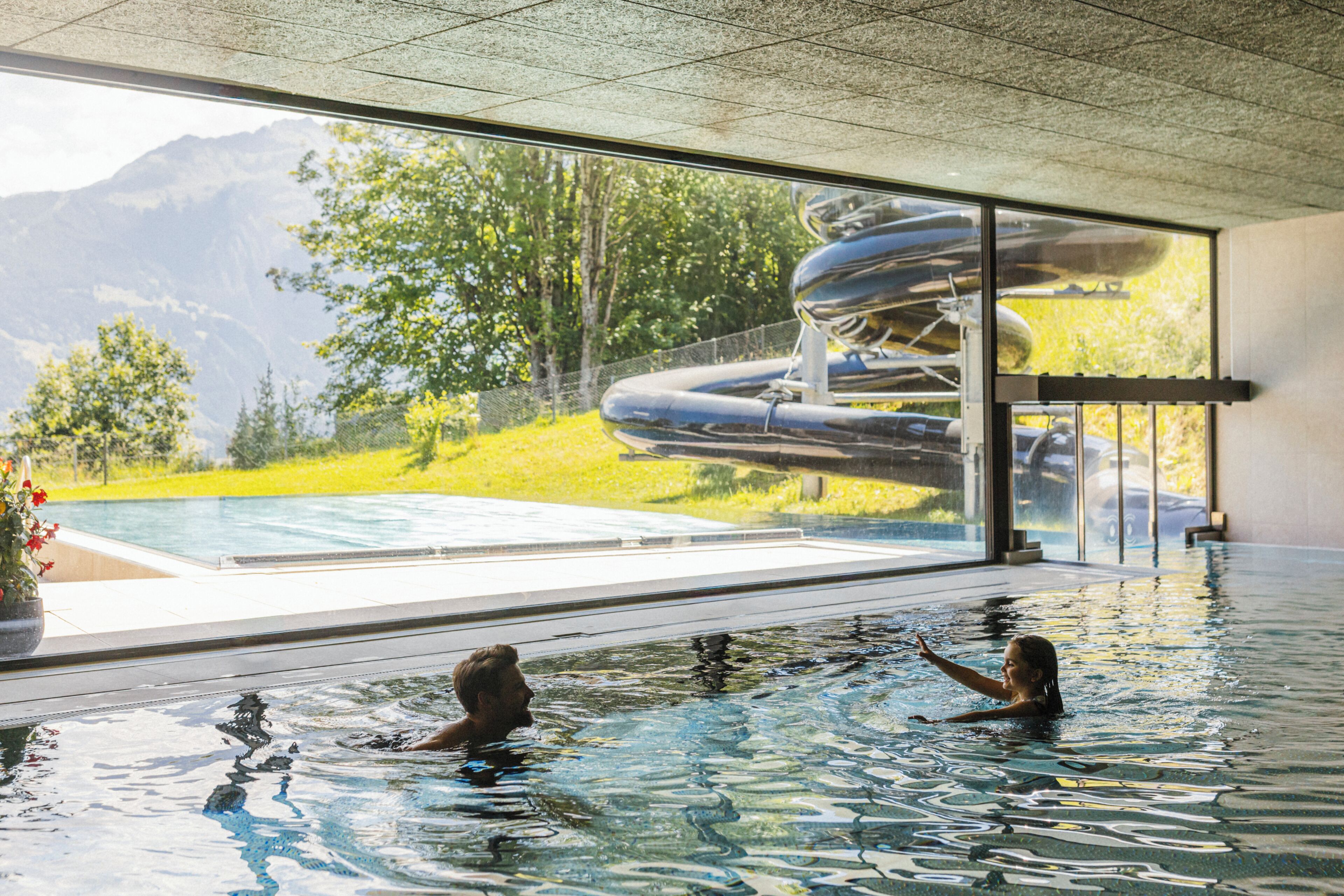 An indoor swimming pool with clear water, a view of a mountainous landscape through large windows, and a slide structure visible outside.
