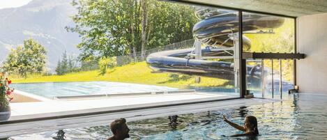 An indoor swimming pool with clear water, a view of a mountainous landscape through large windows, and a slide structure visible outside.