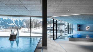 Una piscina interior moderna con paredes de vidrio, zona de sauna y vistas a montañas nevadas.