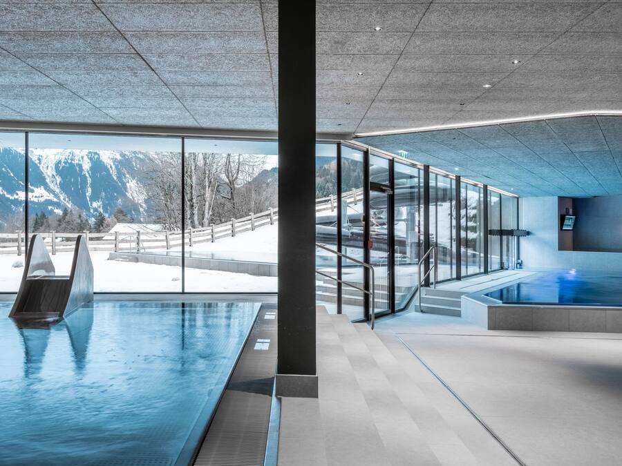 Piscina interior moderna com paredes de vidro, área de sauna e vista para montanhas cobertas de neve.
