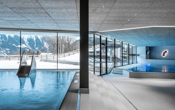 Una piscina interior moderna con paredes de vidrio, zona de sauna y vistas a montañas nevadas.