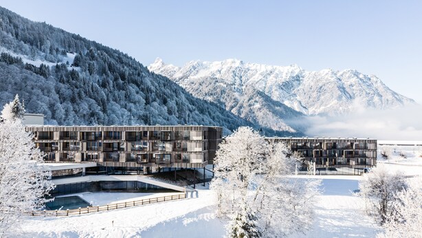 Un edificio moderno con fachada de vidrio y techo plano, rodeado de árboles y montañas cubiertas de nieve.