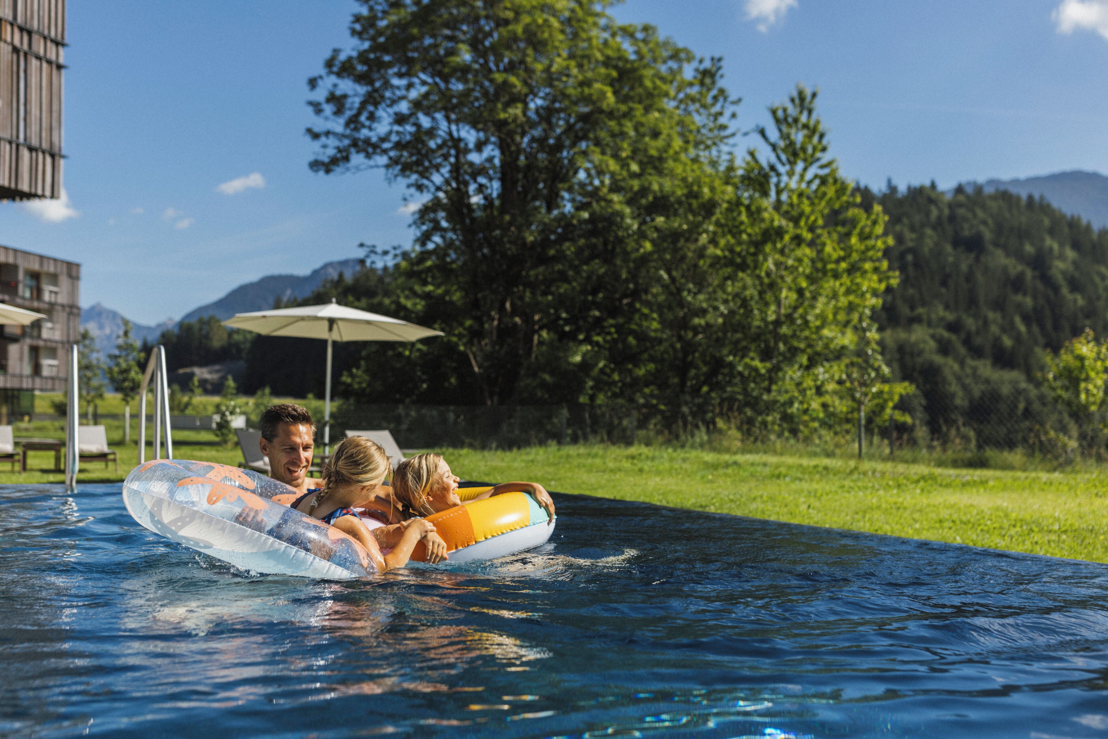 A swimming pool with a couple on a float, surrounded by greenery and a building in the background.