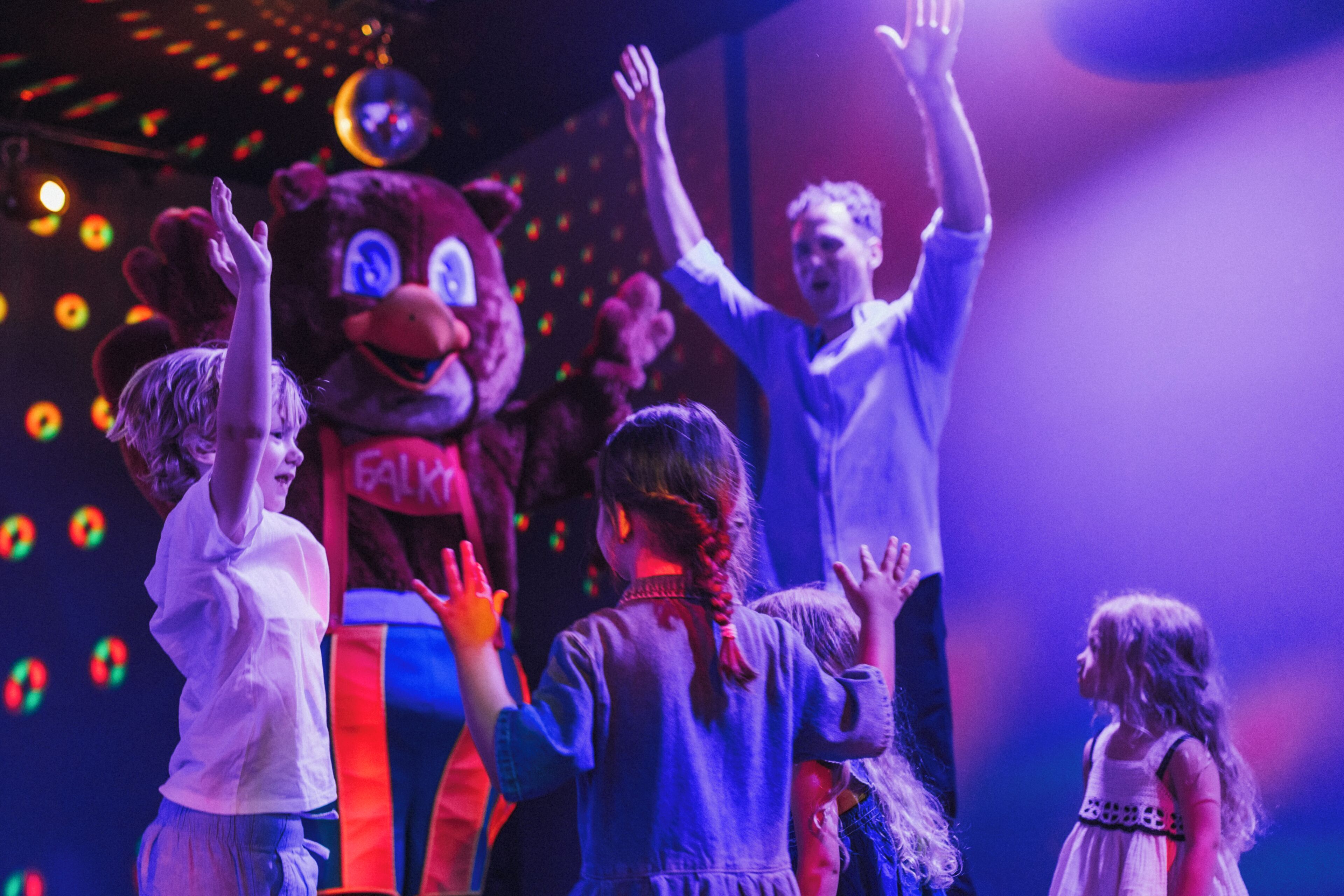 A group of children interacting with a mascot on a stage with colorful lights in the background.