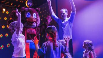 A group of children interacting with a mascot on a stage with colorful lights in the background.