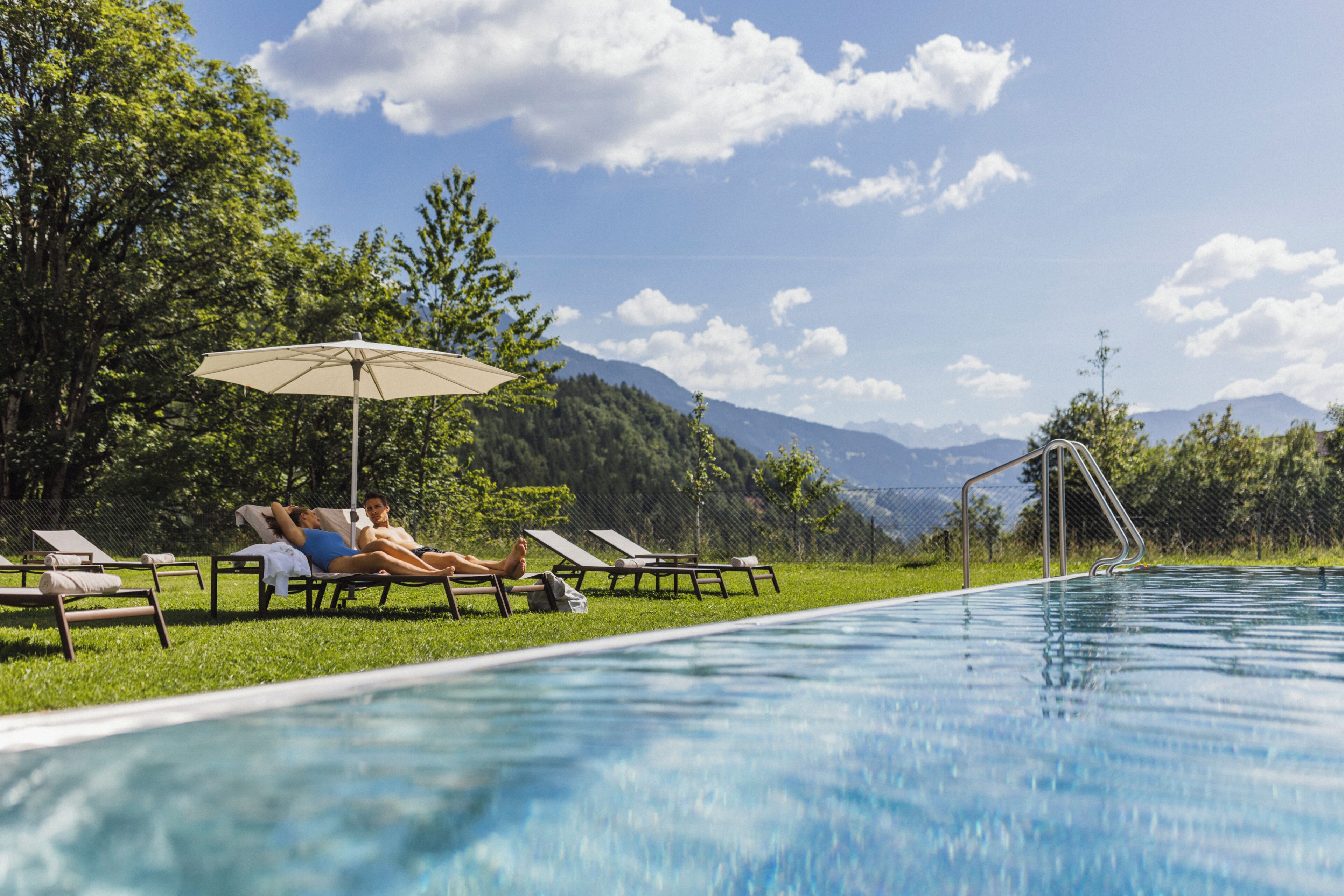 Ein Swimmingpool mit Liegestühlen und einem Sonnenschirm, umgeben von viel Grün und Bergen.
