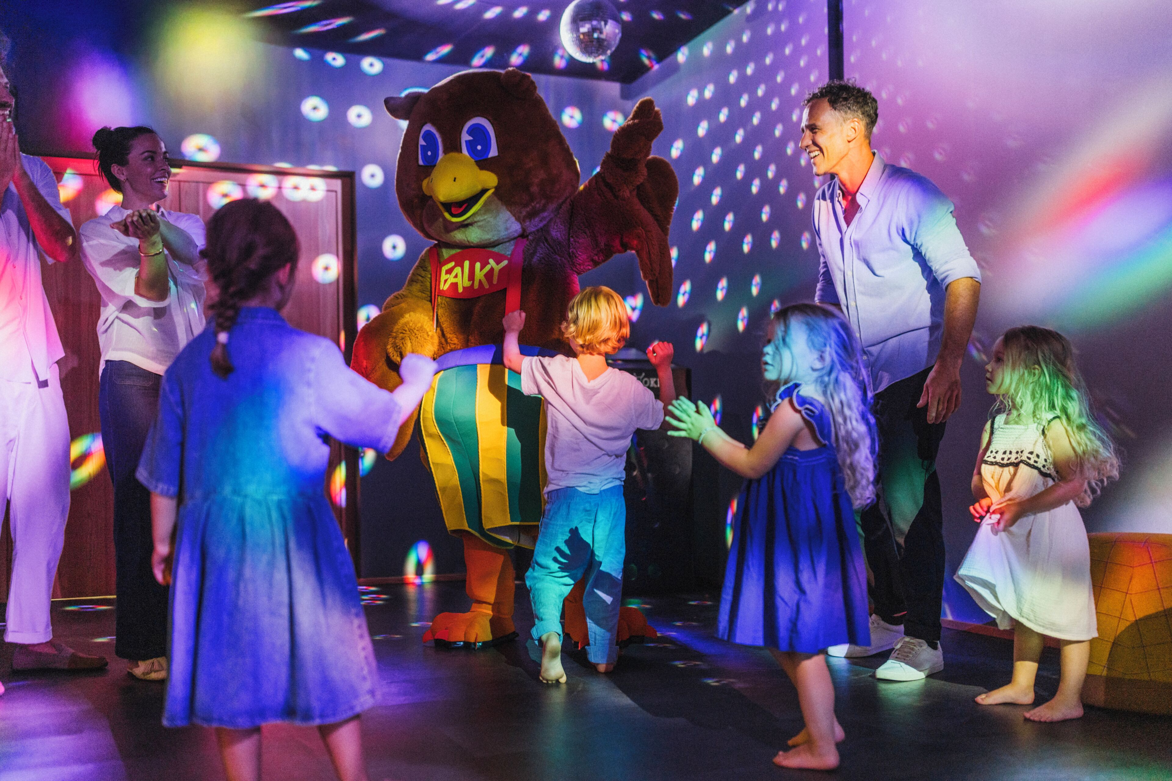 A group of people interacting with a mascot in a room with a patterned wall and disco lights.
