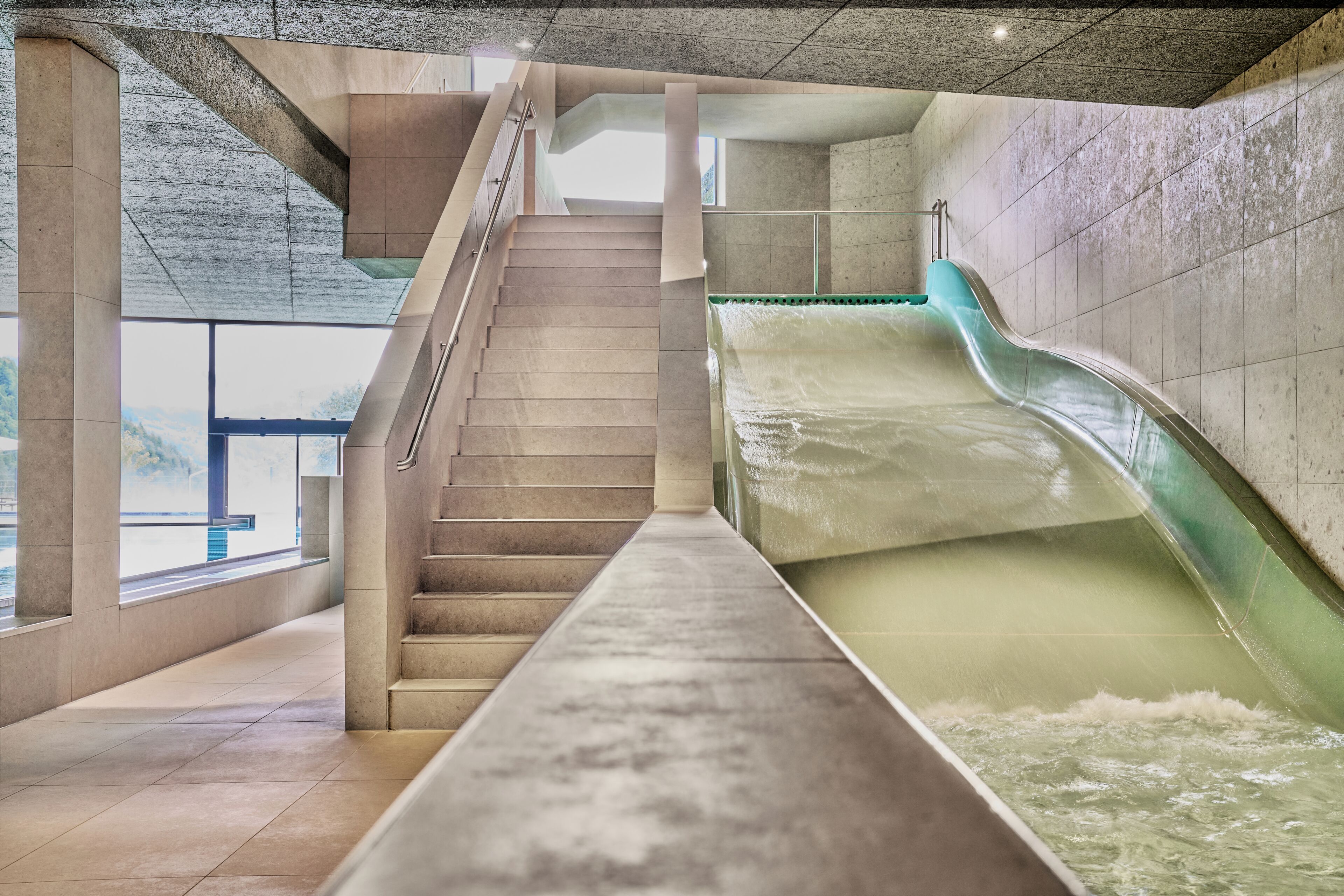Un tobogán de agua moderno en el interior, con un tramo recto y una pendiente suave, rodeado de paredes de piedra y una escalera.