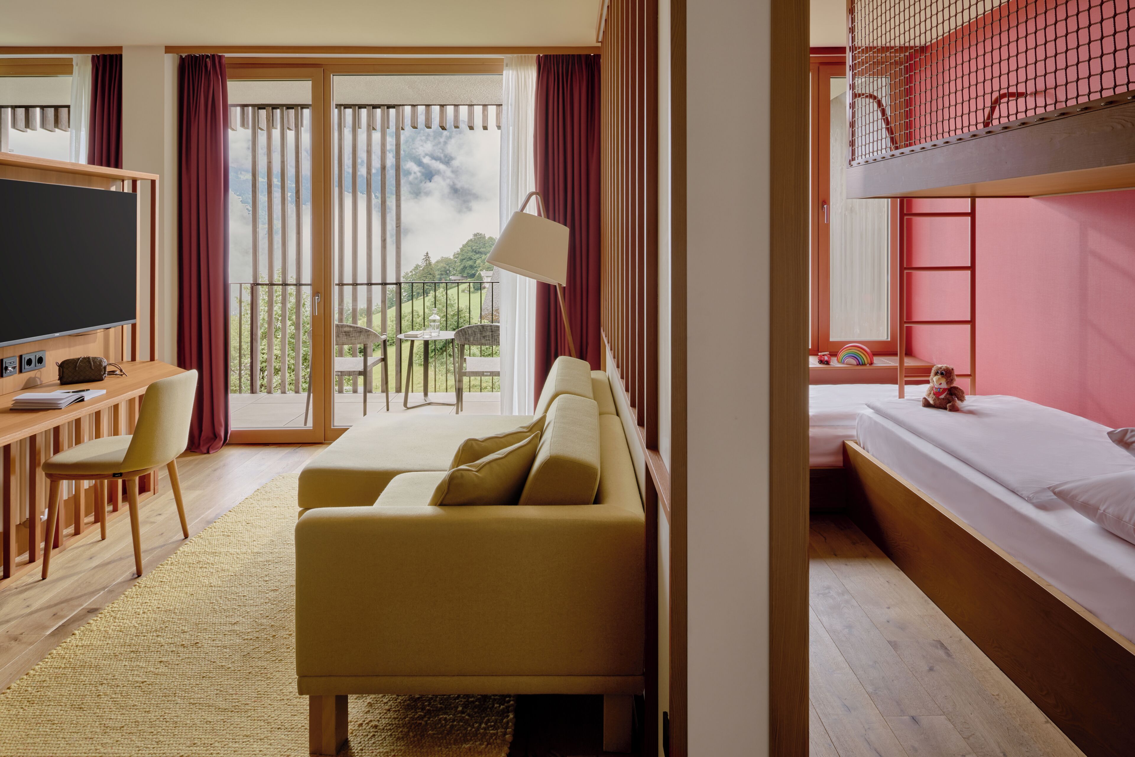 A hotel room with a sofa, a desk with a TV, a balcony with a view of the countryside, and a bunk bed.