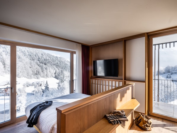Una habitación de hotel moderna con una cama grande, cabecera de madera, un televisor de pantalla plana montado en la pared y un balcón con vistas a un paisaje nevado de montaña.
