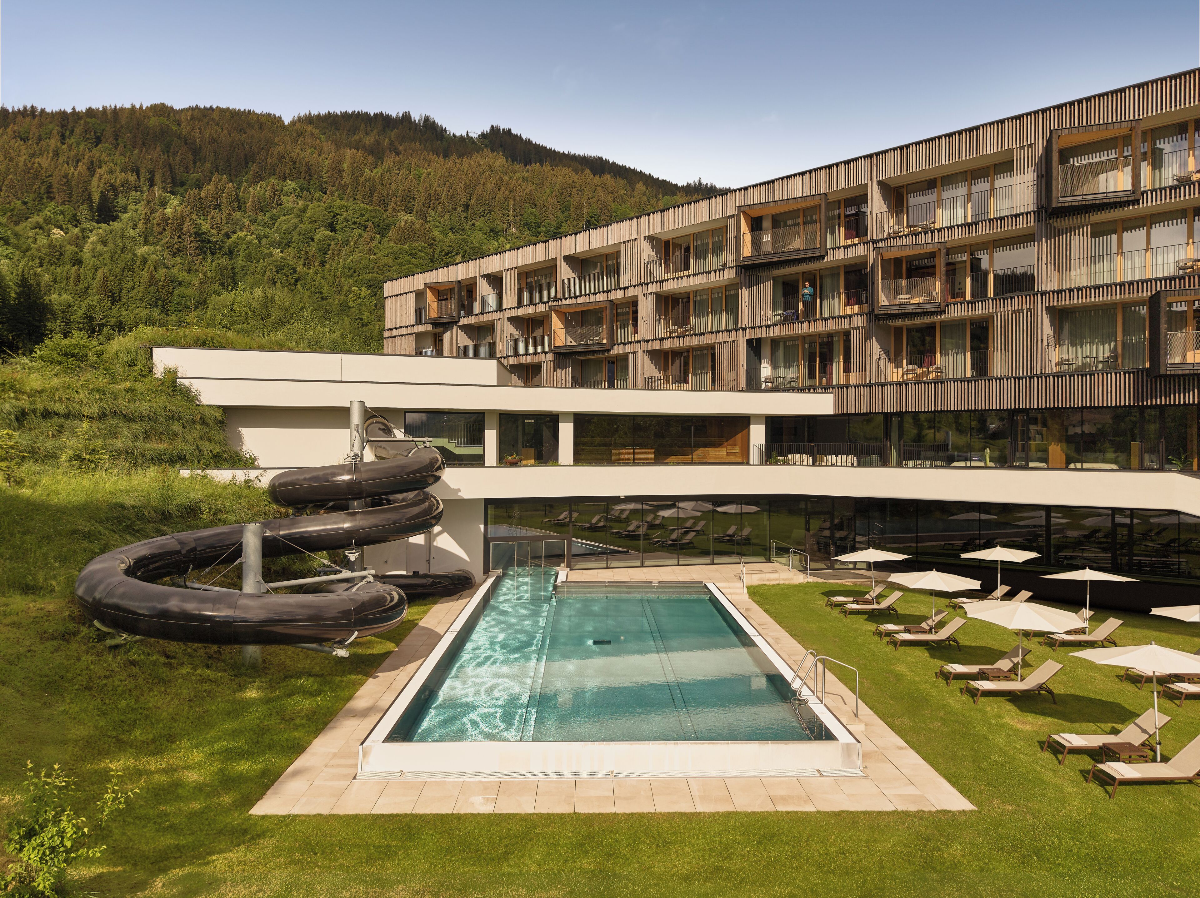 A modern hotel with a pool, outdoor seating, and a view of a forested hill.