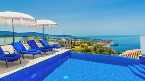 A poolside area with blue lounge chairs and white umbrellas, overlooking a coastal landscape with a clear view of the sea and distant hills. - Villa La Palmera with private pool and fantastic views (Salobreña)
