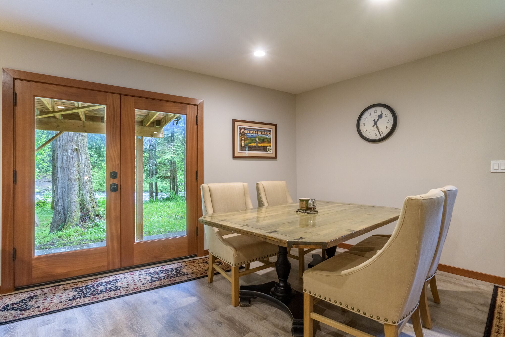 Un coin repas avec une table en bois et quatre chaises, une horloge au mur et une porte donnant sur une zone boisée.