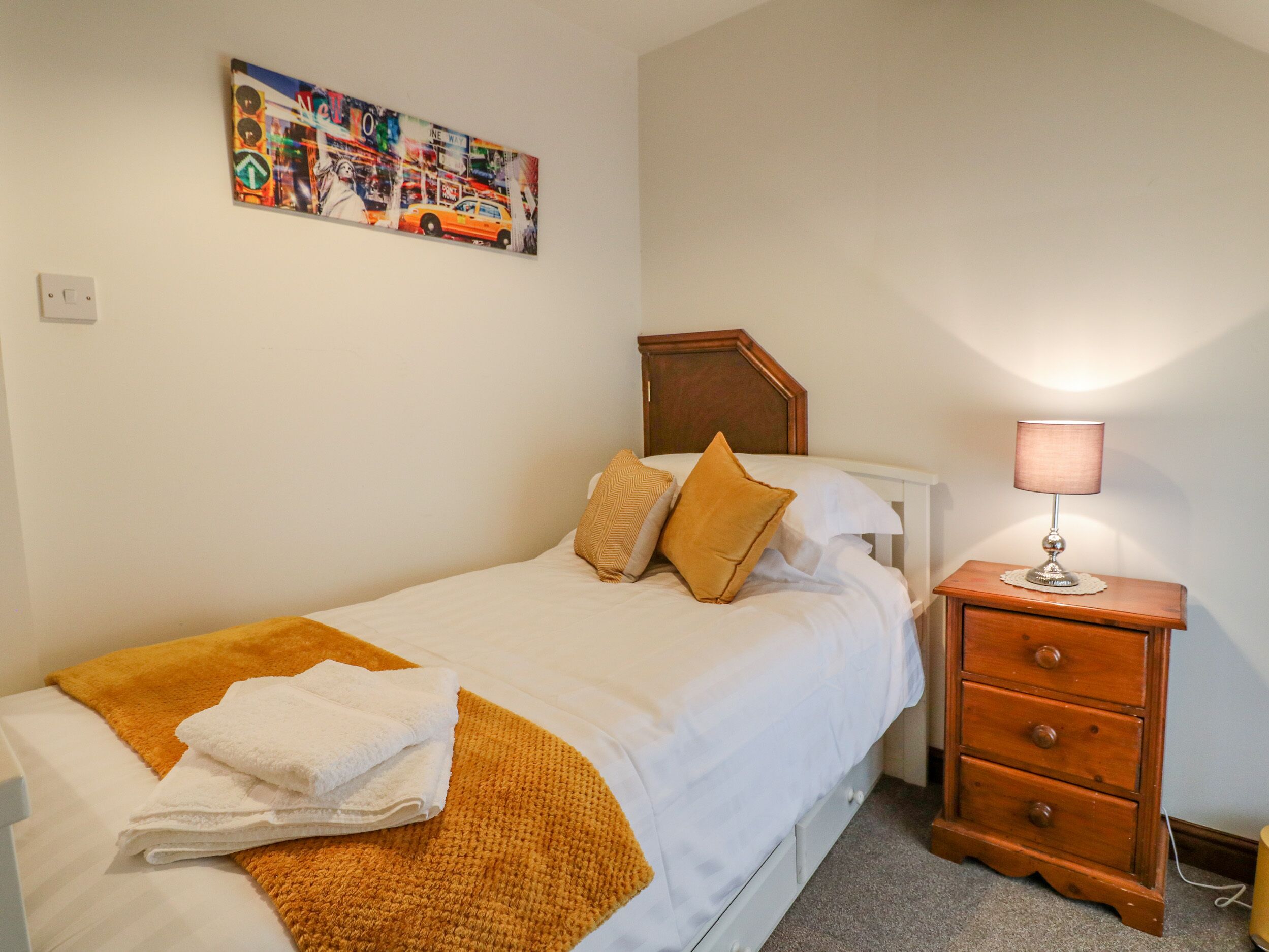 A neatly made bed with white linens, a brown throw blanket, and a folded white towel. There is a wooden nightstand with a lamp and a small drawer. A colorful painting hangs on the wall above the bed.