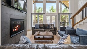 A modern living room with a fireplace, a large window, and a staircase. - Relax and Unwind, Hot Tub Under the Stars, Firepit, Sleeps 8 (broken bow)