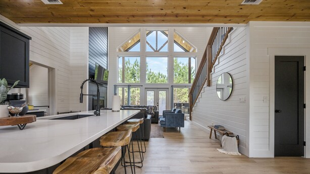 A kitchen with a long counter, bar stools, and a view of the outdoors through large windows. - Relax and Unwind, Hot Tub Under the Stars, Firepit, Sleeps 8 (broken bow)