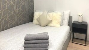 Un lit bien fait, avec une couverture grise et des oreillers, une petite table de chevet noire surmontée d’un vase, et un mur orné d’un motif géométrique.