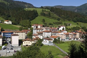 A town with a mix of residential buildings, including multi-story apartments and houses with red-tiled roofs, surrounded by green hills and trees.