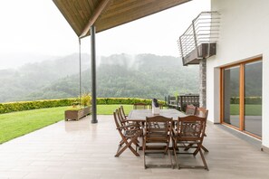 A covered patio with a table and chairs, overlooking a lush garden and distant hills.