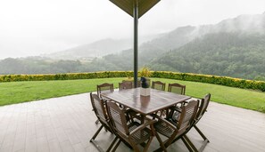 An outdoor patio with a wooden table and chairs, surrounded by a hedge and a view of misty mountains.